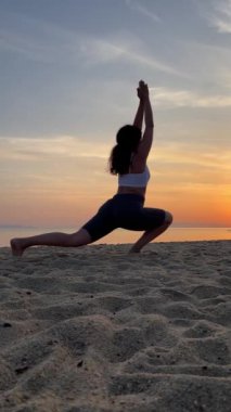 Şafakta kumsalda yoga yapan bir kız, savaşçı pozu.