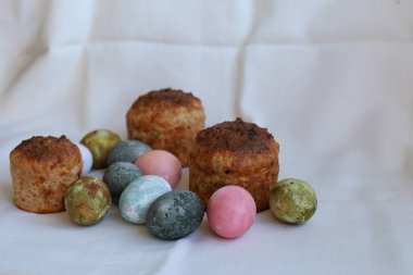 Üç Paskalya pastası ve beyaz masa örtüsü üzerinde renkli yumurtalar.