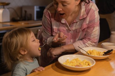 Annesi oğlunu mutfakta çatalla çırpılmış yumurtalarla besliyor.