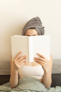 Kafasında havlu olan bir kadın yatağında oturuyor ve yüzüne bir kitap tutuyor.