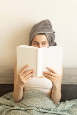 Kafasında havluyla elinde kitap tutan ve başka tarafa bakan bir kadın.