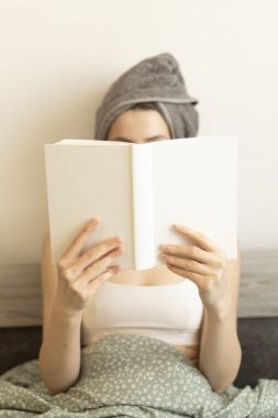 Kafasında havlu olan bir kadın yatakta oturur ve yüzüne bir kitap tutar.