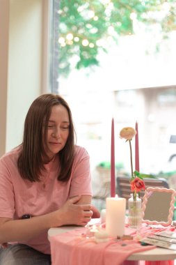 Genç bir kadın, yumuşak süslenmiş pembe bir masada tarot konsültasyonu alıyor, manevi sezgileri ve kişisel berraklığı keşfediyor..