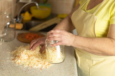 Önlüklü olgun bir kadın sıcak bir ev mutfağında kavanoza lahana karışımı sıkıştırıyor..