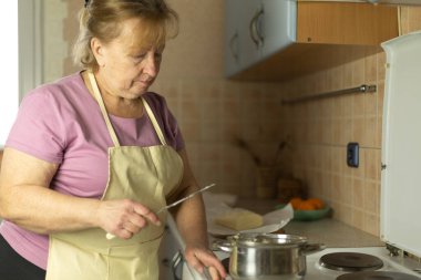 Önlüklü yaşlı bir kadın fırının üzerindeki tencereye tereyağı eklemek için bıçak kullanıyor..