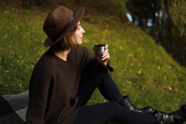 Kadın sakin bir gölün kenarında otururken termos bardağından çay içiyor, açık havada sessiz bir sonbahar akşamının tadını çıkarıyor..