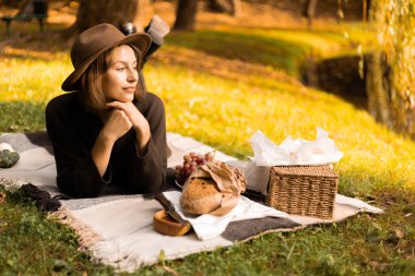Öğleden sonra güneş ışığı huzurlu bir piknik sahnesini aydınlatıyor. Bir kadın bir sepet mevsimlik ikramla uzanırken..