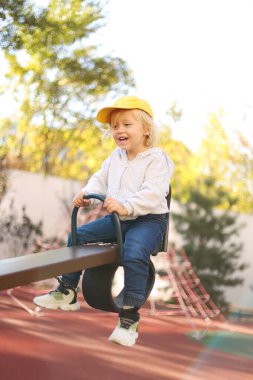 Sarı şapkalı küçük bir çocuk parkta oynarken tahterevallide eğleniyor.