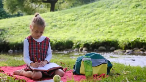 公園の芝生の上に女の子が座って本を書いている 川のそばで自然の中で宿題をしています — ストック動画