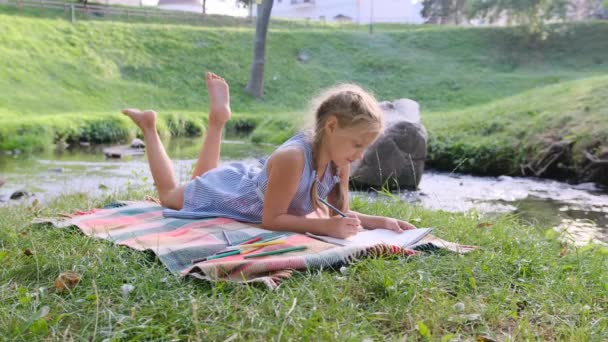 少女がアルバムに鉛筆で描いた川のほとりに横たわっています 子供は晴れた日を楽しんでいます — ストック動画