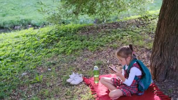 木の下の屋外公園の緑の芝生の上に座って スマートフォンの画面を見て 屈託のない女の子は オンラインゲームをプレイしている — ストック動画