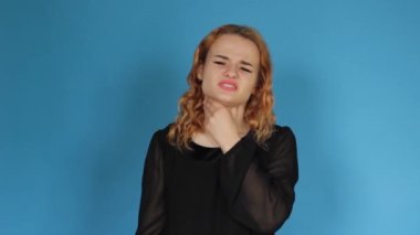 An unhealthy young woman touches her neck, it is difficult to swallow, experiencing discomfort or a painful feeling, isolated on a blue background studio