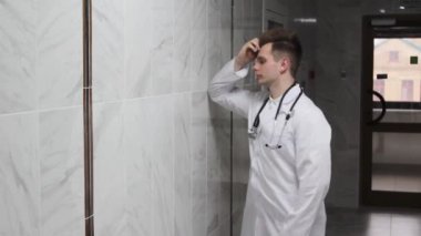 A concerned male doctor leaning on a wall near a corridor in a hospital