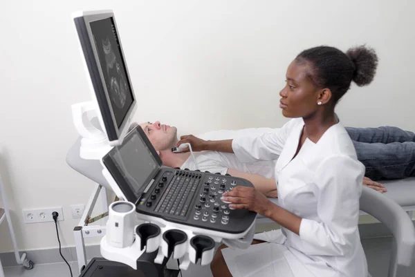 Ultrasound diagnostics of the thyroid gland of a man in the clinic, an African-American doctor launches an ultrasound sensor