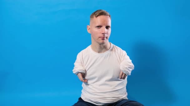 腕と足の先天性病理の白いTシャツを着た若い男が青い背景で微笑む — ストック動画