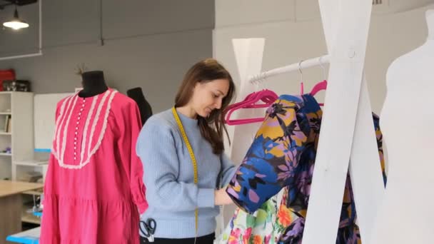 首周り1センチの若いデザイナーの女の子がワークショップでハンガーにぶら下がっている新しいコレクションのドレスを検査します — ストック動画
