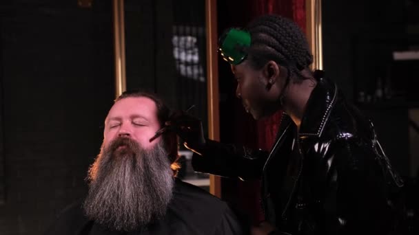 黒いレインコートを着たアフリカ系アメリカ人の女性が床屋で剃刀で男のひげを剃る — ストック動画