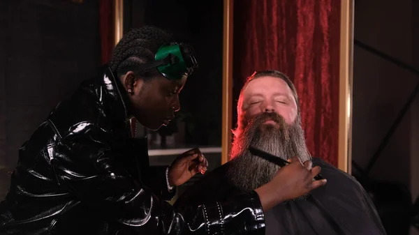 An African-American hairdresser girl combs and cuts a man\'s beard