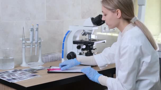 Жіночий Лабораторний Асистент Вивчає Мікроорганізми Переглядаючи Мікроскоп Записує Дані Журналі — стокове відео