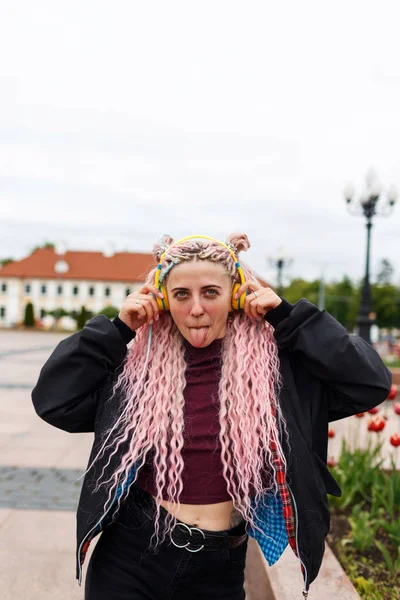 ピンクのドレッドヘアの若い美しい女の子は彼女の舌を示し 街の通りにヘッドフォンで音楽を聴く — ストック写真