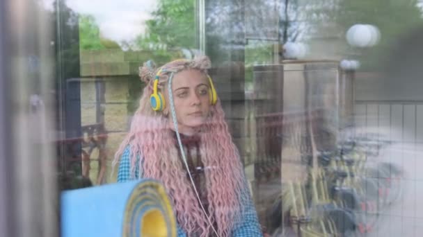 Dziewczyna Długimi Różowymi Włosami Żółtych Słuchawkach Siedzi Szybą Kawiarni Słuchając — Wideo stockowe