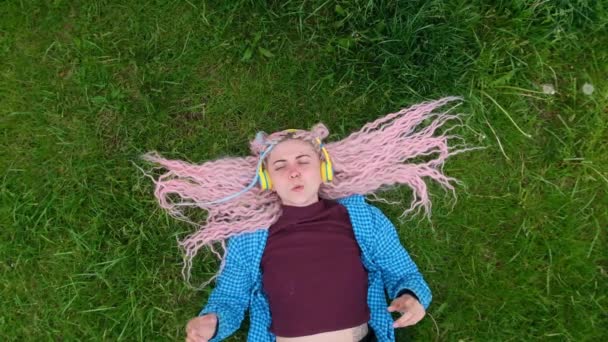 这个女孩很高兴听音乐 在户外放松 在草地上的年轻女子 一头粉色的头发 动作缓慢 — 图库视频影像