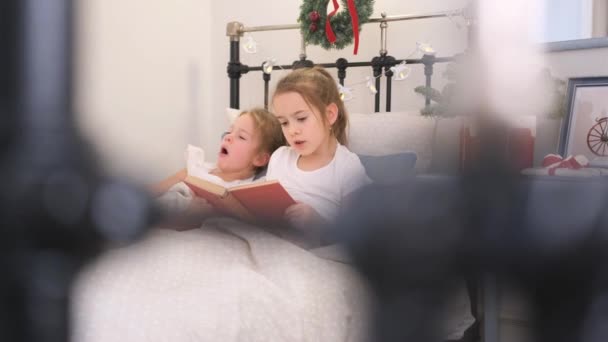 お姉さんは寝る前に妹のために本を読みます 小さな姉妹が同じベッドに横たわっている — ストック動画
