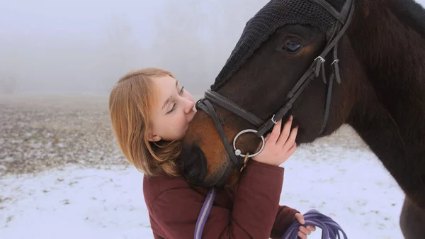 A beautiful teenage girl playfully kisses a brown horse in a foggy field