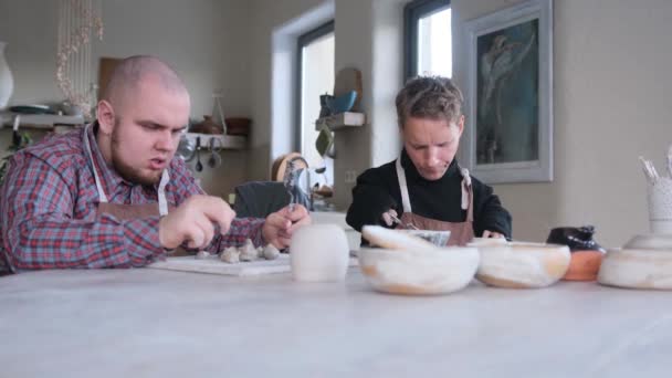 障害のある2人の若者が 大きなテーブルに座っているホームワークショップで粘土板を彫っています — ストック動画