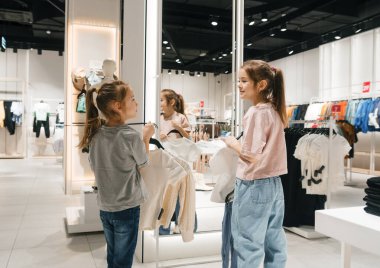 İki kız, gözle görülür şekilde heyecanlı, kalabalık bir mağazada giysi raflarını karıştırıyor, yeni favori kıyafetlerini arıyorlar..