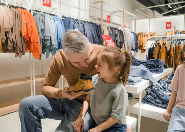 stock image A father playfully engages with his daughter while shopping for shoes in a clothing store, surrounded by racks of clothes and discount signs.