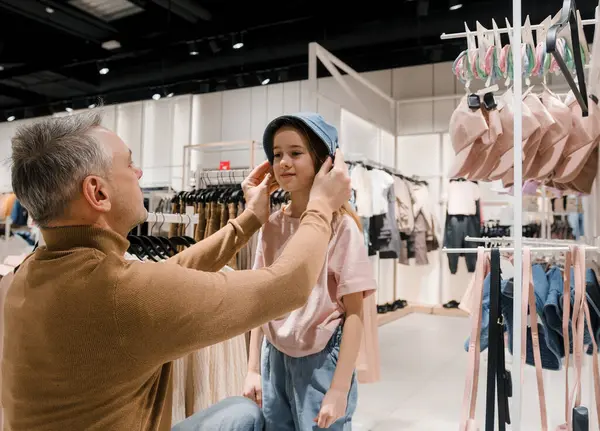 stock image A father adjusts a hat on his daughters head as they browse through clothing racks in a bright, modern store.