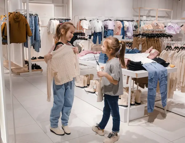 stock image Two young girls excitedly browse and discuss clothing choices in a well-lit childrens fashion store filled with cozy apparel.