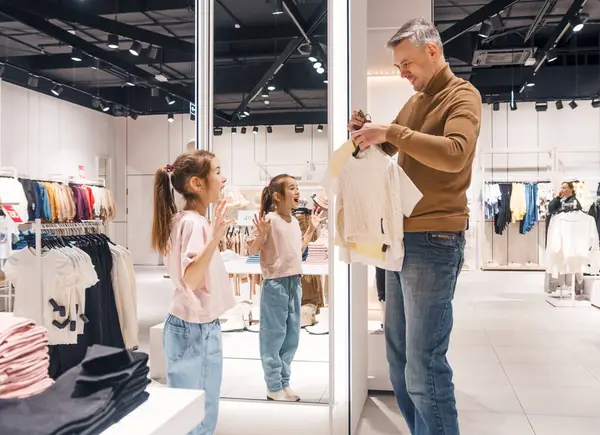 stock image A father helps his young daughter choose clothes in a modern retail store, examining a top held up to her.