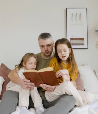 Bir baba yatağa oturur ve iki kızına kitap okur. Kızlar dikkatle dinliyorlar, hikayeden büyülenmişler. Yüzleri merak ve heyecanla dolu, paylaşılan anların neşesini ve hikaye anlatmanın gücünü yansıtıyorlar. Resim ca