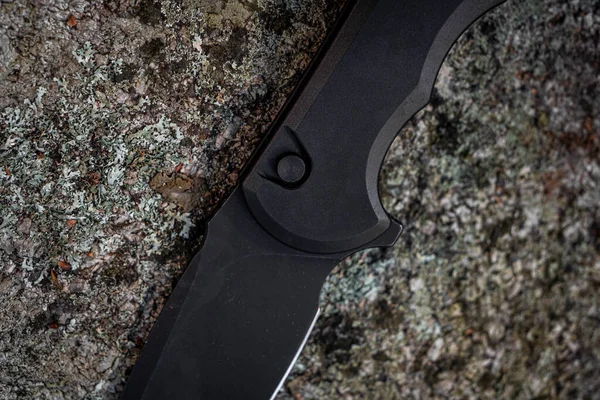 A solid matte black pocket knife sitting on a stone in the wilderness