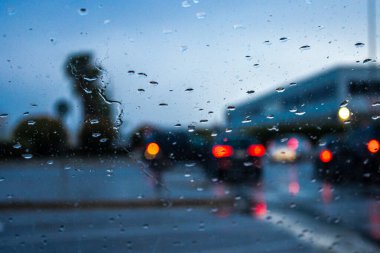 Yağmur mevsiminde yağmur mevsiminde bulutlu gökyüzüne karşı asfalt caddeye ve bina binasına akın ederken Bokeh kırmızı stop lambalarını yansıtan araçların cam manzarasından.