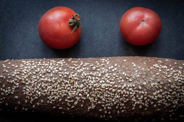 Bu görüntüde iki olgun domates, koyu taş bir yüzeye kurulmuş bir somun susamlı ekmeğin yanında, taze ürünler ve sağlıklı yiyecekler sergiliyor..