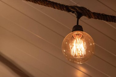  Kalın bir halat armatüründen sarkan parıldayan Edison tarzı bir iplik ampulünün yakın çekimi sıcak ve rahat bir atmosfer yaratıyor. Enerji, dekorasyon, klasik tasarım ve sıcak ışıklandırma ile ilgili kavramlar için idealdir.