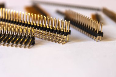 Elektronik devreler ve PCB lehimleme için kullanılan çoklu altın kaplama başlıklı pin bağlantılarının yakın plan görüntüsü. Bileşenler sığ alan derinliğine sahip beyaz bir yüzeye yerleştirilmiş..