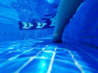  Yüzme havuzunun fayanslı zemininde duran bir çocuğun bacağını gösteren yakın çekim sualtı fotoğrafı. Suyun içinden süzülen güneş ışığı mavi fayanslarda ve deride güzel yansımalar ve desenler yaratır..