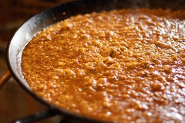 Büyük bir demir tavada pişirilen geleneksel İspanyol paella 'sının yakın çekimi. Sos ve malzemelerin kaynayan dokusu yemeğin kırsal ve ev yapımı doğasını vurgular, sıcak ve davetkar bir aydınlatma mutfak içeriği için mükemmeldir..