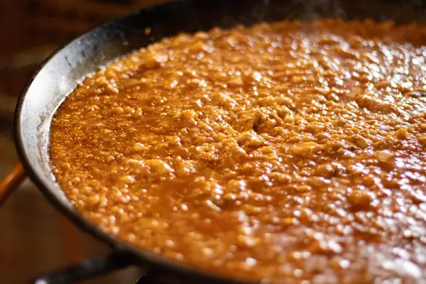 Büyük bir demir tavada pişirilen geleneksel İspanyol paella 'sının yakın çekimi. Sos ve malzemelerin kaynayan dokusu yemeğin kırsal ve ev yapımı doğasını vurgular, sıcak ve davetkar bir aydınlatma mutfak içeriği için mükemmeldir..