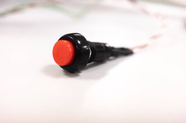 Siyah kaplamalı kırmızı düğmeli makro çekim, temiz beyaz bir yüzey üzerinde duruyor. Görüntü, genellikle elektronik devreler, DIY projeleri ve endüstriyel kontrol sistemlerinde kullanılan düğmenin dokunsal doğasını vurgular.