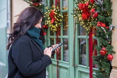 Şenlikli Noel dekorasyonuna sahip akıllı telefon kullanan mutlu bir kadın. Noel, Yeni Yıl ve tatillerde turizm kavramı.