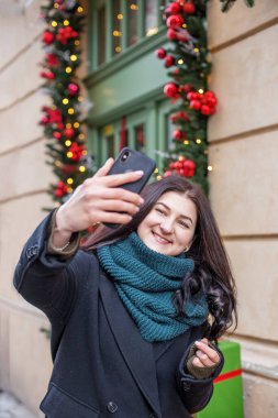 Şenlikli Noel dekorasyonuna sahip akıllı telefon kullanan mutlu bir kadın. Noel, Yeni Yıl ve tatillerde turizm kavramı