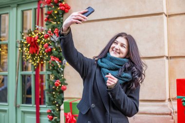 Şenlikli Noel dekorasyonuna sahip akıllı telefon kullanan mutlu bir kadın. Noel, Yeni Yıl ve tatillerde turizm kavramı