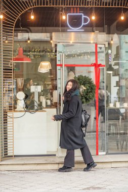 Siyah paltolu genç bir kadın şehirde yürüyor.