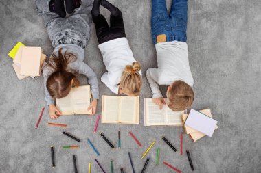 Bir grup çocuk yerde yatarken kitap okuyor ve kalemlerle çizim yapıyorlar. Aile kavramı, eğitim veya eğlence.