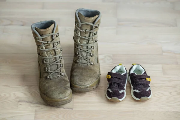 old worn military boots and children\'s sports shoes on wooden floor. Concept of military father and family.